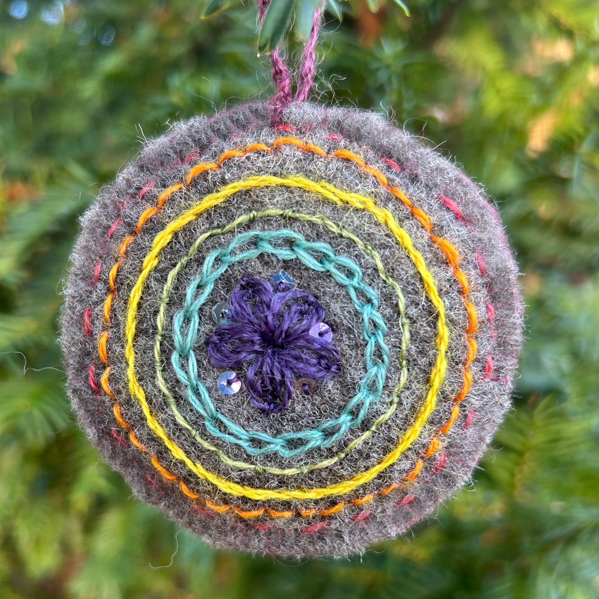 Decorative wool felt embroidery with concentric circles and a purple flower in the center, hanging against a green foliage background.
