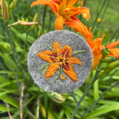 Embroidered day lily design on a textured gray surface with orange daylily in the background