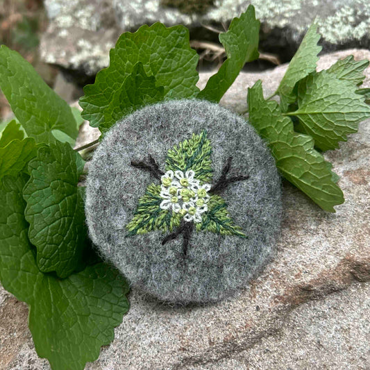 Embroidered Garlic Mustard motif on grey wool felt, surrounded by garlic mustard leaves on stone