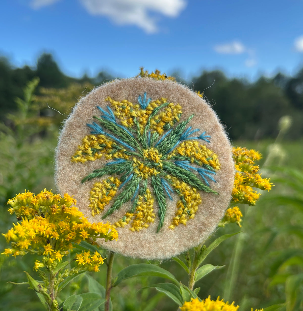 Beige wool felt from local wool, embroidered with a design of goldenrod with green, blue and yellow wool embroidery floss in a field of goldenrod. 